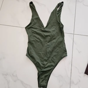 Grön v-ringad bodysuit - Snygg grön bodysuit med djup v-ringning och breda axelband. Modellen är ärmlös och har en tight passform som framhäver siluetten. Perfekt att styla med jeans eller kjol för en cool look.