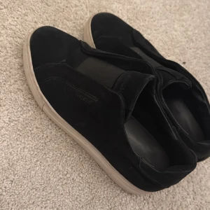 Svarta slip-on sneakers från ARIGATO - Svarta sneakers från ARIGATO med slip-on design och bred elastisk panel över foten. Ovandelen är i mocka och sulan är vit och platt. Snygg minimalistisk look som funkar till jeans eller dressade byxor. Rund tå och diskret logga på sidan.