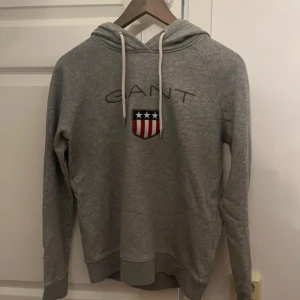 Grå hoodie från GANT - Snygg grå hoodie från GANT med broderat emblem och text på bröstet. Tröjan har huva med vita snören och långa ärmar. Tillverkad i mjuk bomullsmix, perfekt för chill dagar. Klassisk passform och stilren look som funkar året runt.