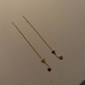 Guldiga örhängen med hjärtan - Säljer ett par tunna, långa örhängen i guldton med två små hjärtan på varje kedja. De är minimalistiska och ger en söt touch till din stil. Perfekta för dig som gillar enkla men ändå unika smycken.