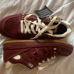 Vinröda Converse One Star sneakers - Säljer ett par Converse One Star sneakers i vinrött mocka med vita kontrastsömmar och detaljer. Skorna har vit snörning, gummisula i naturfärg och klassisk stjärnlogga på sidan. Perfekt för dig som vill sticka ut med en retro vibe.