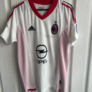 AC Milan Kaka Champions League tröja - Säljer en AC Milan fotbollströja från Champions League-finalen 2003 med Kaka och nummer 22 på ryggen. Tröjan är vit med röda detaljer, svarta Adidas-loggan och Opel-sponsor på bröstet. V-ringad krage och korta ärmar, tillverkad i lätt polyester. Perfekt för dig som älskar retro och fotbollshistoria.