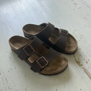 Birkenstock sandaler brun skinn - Bruna Birkenstock sandaler med två breda remmar i skinn och justerbara kopparfärgade spännen. Sval korksula som formar sig efter foten och ger grym komfort. Perfekt för chill dagar och enkel att slänga på sig. Klassisk och tidlös design.
