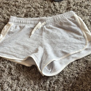 Grå mjuka shorts från H&M Basic - Superbekväma grå shorts från H&M Basic med vit kant och snörning i midjan. Perfekta för chill dagar eller träning. Tillverkade i mjukt bomullsmaterial med loose passform och elastisk midja för extra komfort.