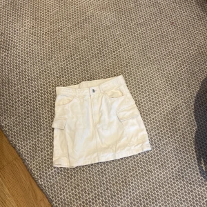 Beige kortkjol/ jeanskjol med fickor från H&M - Snygg beige kortkjol från H&M med praktiska fickor på sidorna och klassisk knappstängning fram. Kjolen har en rak passform och är gjord i mjuk bomull, perfekt för en chill och trendig look. Passar dig som gillar enkel och clean stil.