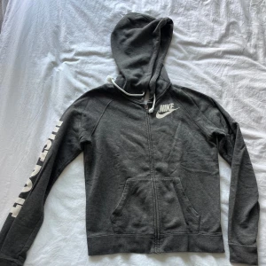 Grå hoodie från Nike med dragkedja - Snygg grå hoodie från Nike med vit logga på bröstet och tryck på ärmen. Tröjan har huva med vita snören, praktiska fickor framtill och dragkedja hela vägen. Perfekt för chill eller träning, riktigt skön och stilren design.