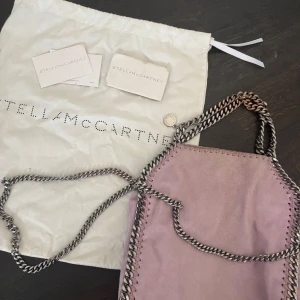 Ljuslila/rosa Stella McCartney väska - Säljer min väldigt välanvända Stella McCartney väska som jag köpte för ungefär ett år sedan här på Plick. Som man kan se så har väskans kedjor börjat tappa färg och mockat är lite smutsigt. Väskans kedjor var redan lite ”rostiga”  när jag köpte väskan och en liten defekt på innersidan av väskan fanns. Jag anser att väskan går ju såklart fortfarande att använda och mockan kan man behandla om man vill💕Dustbag och äkthetsbevis ingår! (Pris går absolut att diskutera☺️)
