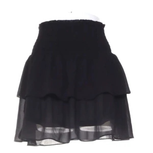 Volangkjol från Chelsea - Säljer en svart kortkjol med bred smockad resår i midjan och dubbla volanger. Kjolen är gjord i ett tunt, lätt och transparent tyg som ger en luftig känsla. Perfekt för dig som gillar en feminin och trendig stil.