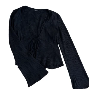 Svart blus med knytning - Snygg svart blus från Gina Tricot med v-ringning och knytning framtill. Blusen har långa ärmar och ett plisserat tyg som ger en elegant look. Perfekt för att höja din stil till nästa nivå.