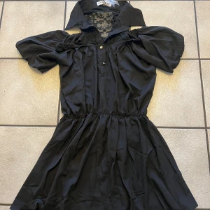 Svart klänning med spetsdetaljer - Snygg svart klänning med spetsdetaljer vid halsen och en elastisk midja. Klänningen har korta puffärmar och en krage som ger en unik look. Perfekt för att sticka ut med stil!