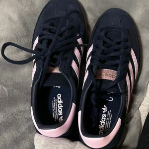 Mörkblåa och rosa sneakers från Adidas - Snygga mörkblåa sneakers från Adidas med rosa ränder och detaljer. Skorna har klassisk snörning och en bekväm passform. Perfekta för en stilren look.