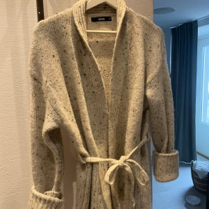 Beige kofta från BikBok - Mysig beige kofta från BikBok med knytband i midjan. Koftan har en naturlig färg med små mörka detaljer i tyget. Använd en gång.