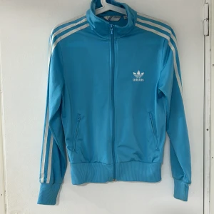 Blå träningsjacka från Adidas - Säljer en snygg blå träningsjacka från Adidas som är använd fåtal gånger (köpte secondhand) med klassiska vita ränder längs ärmarna och en stor logga på ryggen. Jackan har dragkedja framtill och två fickor med dragkedjor. Perfekt för en sportig look eller avslappnad stil. Pris kan diskuteras vid snabb affär.