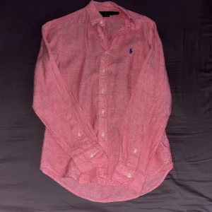 Rosa linneskjorta med broderad logga - Snygg rosa skjorta i linne från Ralph Lauren med en broderad blå logga på bröstet. Skjortan har långa ärmar och knappar framtill. 