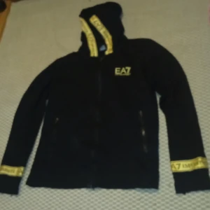 Svart hoodie från EA7 - Snygg svart hoodie från EA7 med gulddetaljer och dragkedja. Den har en huva med guldfärgad text och matchande detaljer på ärmsluten. Perfekt för en sportig look eller avslappnad stil.