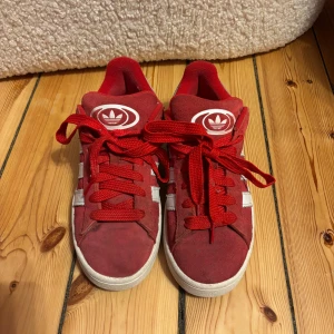 Röda Adidas sneakers barn  - Snygga röda Adidas sneakers med vita detaljer och klassiska tre ränder på sidan. Skorna har snörning och en bekväm sula. Perfekta för en sportig look.