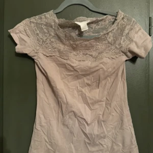 Beige topp med spetsdetaljer - Säljer en söt beige topp med korta ärmar och vacker spetsdetalj runt halsringningen. Perfekt för en stilren och elegant look. Toppen är i ett mjukt och bekvämt material.