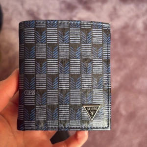 Mönstrad plånbok från Guess - Snygg plånbok från Guess med ett stilrent rutigt mönster i svart och blått. Plånboken har flera kortfack och en sedelficka. Perfekt för att hålla ordning på dina kort och kontanter. Guess-logotypen syns både på utsidan och insidan.