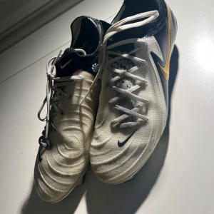 Nike Phantom GX fotbollsskor - Säljer ett par Nike Phantom GX fotbollsskor i vitt och svart med gulddetaljer. Skorna har snörning och är designade för optimal prestanda på planen. Perfekta för dig som vill ha stil och funktion i ett.