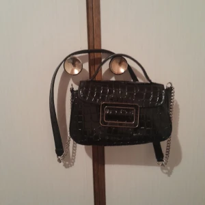 Zara axelväska med kedjedetaljer - Snygg svart Zara axelväska med krokodilpräglat mönster och kedjedetaljer. Väskan har en dekorativ spänne på framsidan och ett praktiskt handtag. Perfekt för att ge din outfit en elegant touch.