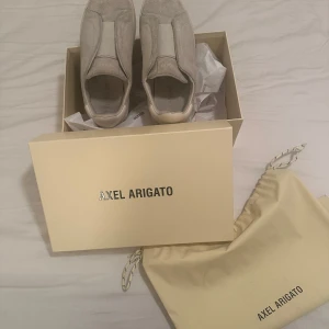 Beige sneakers från Axel Arigato - Snygga beige sneakers från Axel Arigato i mocka. Skorna har en stilren design utan snörning och en robust sula med märkets logotyp. Perfekta för en avslappnad och trendig look.köpta för 2800 i våras. Kommer med låda, dustbag