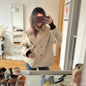 Beige trenchcoat - Säljer en super populär kort beige trenchcoat med dubbelknäppning och perfekt oversize fit på mig som oftast bär xs!!