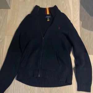 Svart kofta från Polo Ralph Lauren - Säljer en stilren svart kofta från Polo Ralph Lauren med dragkedja framtill. Den har ribbade muddar och en liten röd logotyp på bröstet. Perfekt för kyligare dagar.