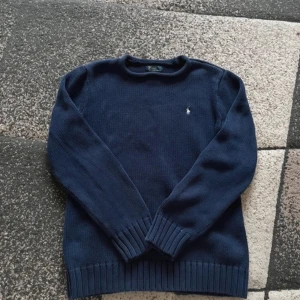 Mörkblå stickad tröja från Ralph Lauren - Säljer en stilren mörkblå stickad tröja från Ralph Lauren med en liten broderad logga på bröstet. Tröjan har långa ärmar och ribbstickade muddar vid ärmslut och nederkant. Perfekt för kyliga dagar!