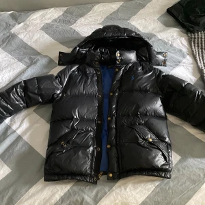 Svart dunjacka från Polo - Säljer en svart dunjacka från Polo med blått foder. Jackan har en huva och stängs med dragkedja och knappar. Perfekt för kyliga dagar! Äkta jacka!!!💙