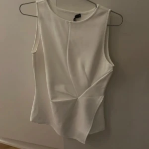 Vit ärmlös topp från BikBok - Säljer en stilren vit ärmlös topp från BikBok. Toppen har en elegant design med asymmetriska sömmar framtill som ger en modern touch. Perfekt för att matcha med både jeans och kjol. Bara använd en gång
