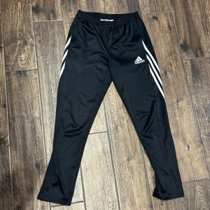 Svarta träningsbyxor från Adidas - Snygga svarta träningsbyxor från Adidas med de klassiska tre vita ränderna längs sidorna. Byxorna har en elastisk midja och är perfekta för sport och träning. Adidas-loggan är broderad på framsidan. Defekter finns och syns på bild