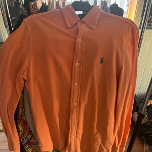 Orange långärmad skjorta från Ralph Lauren - Snygg orange långärmad skjorta från Ralph Lauren med klassisk krage och knappar framtill. Den har en broderad logotyp på bröstet och är tillverkad i ett lätt och bekvämt material.
