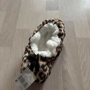 Leopardmönstrade tofflor med rosett - Mysiga tofflor med leopardmönster och en söt svart rosett. Insidan är fodrad med mjukt vitt material för extra komfort. Perfekta för att hålla fötterna varma och stiliga hemma. Storlek 36-38 helt nya!