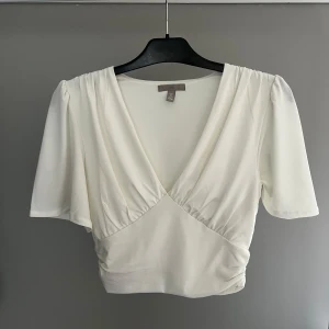Vit v-ringad topp från H&M - Säljer en stilren vit topp från H&M med v-ringning och korta ärmar. Toppen har en elegant drapering framtill och är perfekt för en chic look. Passar bra till både jeans och kjol.