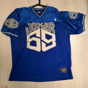 Joker brand jersey  - Säljer en blå jersey från joker brand med stort Joker 69-tryck i silver på både fram- och baksidan. Tröjan har korta ärmar och en v-ringad krage med grå detaljer. Perfekt för en unik och sportig stil!