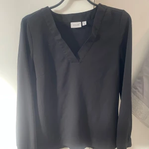 Svart blus från Vila - Snygg svart blus från Vila med v-ringning och långa ärmar. Perfekt för en stilren look. Tillverkad i ett mjukt material som ger en bekväm passform.