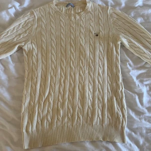 Vit stickad tröja från Gant - Säljer en stilren vit stickad tröja från Gant med klassiskt flätmönster. Tröjan har långa ärmar och rund halsringning, perfekt för kyligare dagar. En liten logotyp på bröstet ger en extra touch av stil.