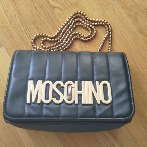 Svart axelväska från Moschino - Snygg svart axelväska från Moschino med quiltad design och en elegant kedjerem i guld. Väskan har en stor logotyp på framsidan som ger en lyxig känsla. Perfekt för att lyfta vilken outfit som helst.