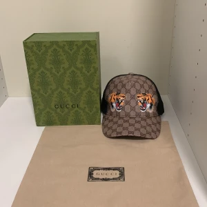 Gucci keps med tigerbroderi - Snygg Gucci keps med ikoniskt GG-mönster och broderade tigrar framtill. Kepsen har en justerbar rem baktill med klassiska Gucci-färger. Perfekt för att ge din outfit en lyxig touch. Priset kan diskuteras, kepsen är äkta dock finns det inget kvitto. Kepsen är helt oanvänd