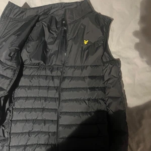 Svart dunväst med dragkedja - Snygg svart dunväst från Lyle & Scott med dragkedja och deras ikoniska gula logga på bröstet. Perfekt för lager-på-lager under kyliga dagar. Västen har en stilren design med quiltade paneler.