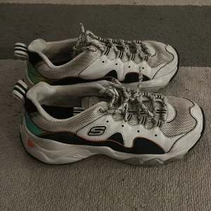 Vita sneakers från Skechers - Snygga vita sneakers från Skechers med svarta och gröna detaljer. Skorna har snörning och en bekväm sula, perfekt för vardagsbruk. De har en sportig design med märkets logotyp på sidan.