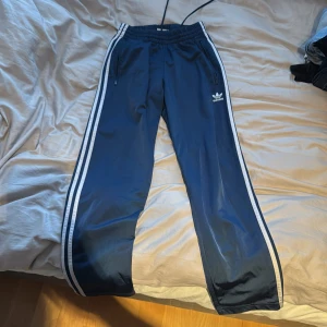 Blå träningsbyxor från Adidas - Snygga blå träningsbyxor från Adidas med vita ränder längs sidorna. Byxorna har en elastisk midja och dragkedjor vid fickorna. Perfekta för en sportig look.