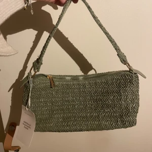 Grön axelväska med dragkedja  - Snygg grön axelväska i flätat material med dragkedja upptill. Perfekt för att ge en naturlig touch till din outfit. Väskan har en enkel design med en bekväm axelrem. 