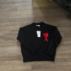 Svart tröja från Ami Paris - Snygg svart tröja från Ami Paris med ett rött hjärta och bokstaven A broderat på bröstet. Tröjan har långa ärmar och ribbade muddar vid ärmslut och nederkant. Perfekt för en stilren look.