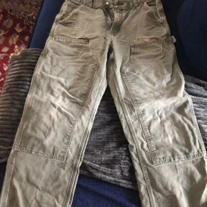 Gröna cargobyxor från Carhartt - Säljer ett par gröna double knee byxor från Carhartt i bomull. Byxorna har flera praktiska fickor och en dragkedja framtill. Perfekta för en avslappnad stil med en loose passform. Knappen på byxorna har lossnat så jag har satt ditt en temporär lösning