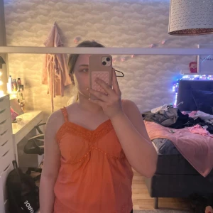 Korall linne med spetsdetaljer - Säljer ett snyggt korall linne med spetsdetaljer och justerbara axelband. Toppen har en vacker broderad design och pärldetaljer framtill. Perfekt för sommardagar! (Linnet ser mycket orangeare ut på de tre första bilderna men se sista bilden för en lite mer noggran bild på färgen).