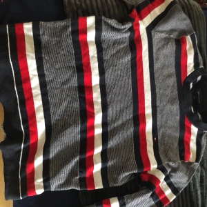 Randig tröja från Tommy Hilfiger - Säljer en randig tröja från Tommy Hilfiger i svart, vit och röd. Tröjan har långa ärmar och ett klassiskt mönster med breda ränder. Tröjan har sönder vid ena armbågen och behöver sys samt vid kragen hänger det en textil bit 