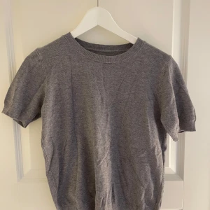 Grå kortärmad tröja - Säljer en stilren grå tröja med korta ärmar. Tröjan har en rund halsringning och ribbade kanter vid ärmslut och nederkant. Perfekt för en avslappnad look. Nyskick.