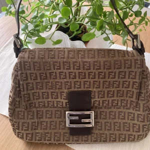 Vintage Brun axelväska från Fendi - Säljer en snygg brun axelväska från Fendi med det ikoniska FF-mönstret. Väskan har en stilren design med ett metallspänne framtill och justerbar axelrem. Perfekt för att ge din outfit en lyxig touch.