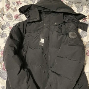 Svart dunjacka från Canada Goose - Säljer en svart dunjacka från Canada Goose med pälskantad huva och dragkedja. Jackan har knappar och en broderad logotyp på ärmen. Perfekt för kalla vinterdagar.